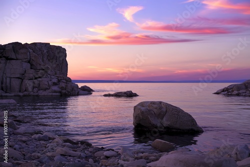 A rocky shoreline with a calm lake or ocean in the foreground, and a colorful sunset sky wi