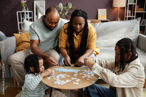 Family gathered around wooden table solving puzzle, focusing on pieces and working together. Living space is warm and inviting, with plants and lamps adding ambiance photo