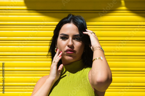 Young Spanish woman, brunette and beautiful with green dress on yellow background touches her hair and face and looks at the camera in a daring and shameless way. Beauty and fashion concept.