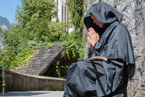 An old monk in a black cassock and carrying a staff is praying with his hands folded and leaning against the wall. photo