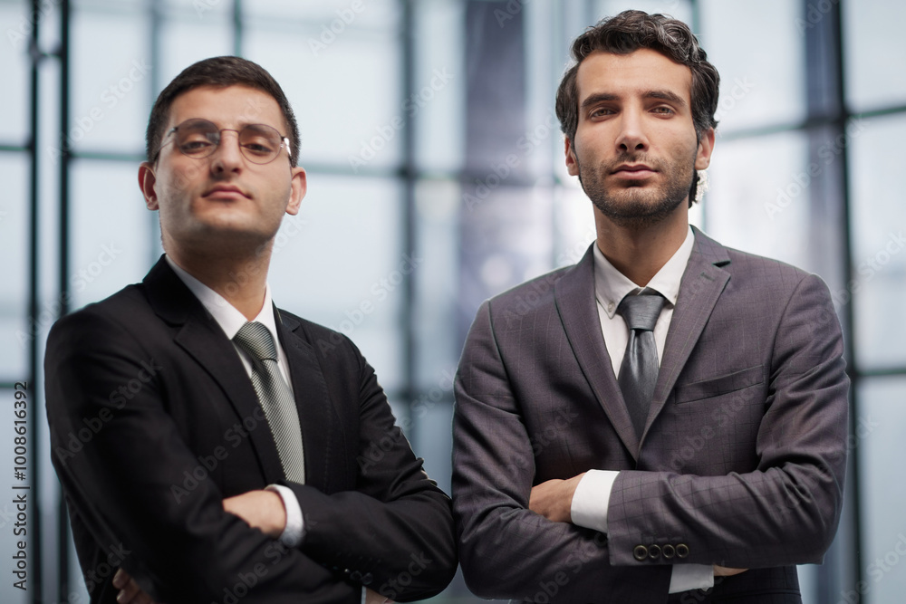 Two serious young businessmen standing with arms crossed in office