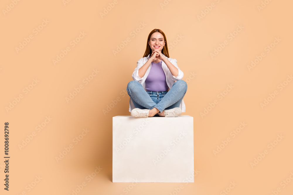 Full size photo of intelligent positive woman wear white cardigan jeans sit on cube arms under chin isolated on beige color background Full size photo of intelligent positive woman wear white cardigan jeans sit on cube arms under chin isolated on beige color background