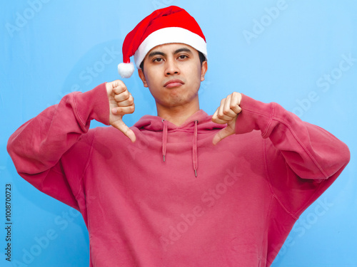 Disappointed young Asian man in a red hoodie and Santa hat giving a thumbs down at the camera against a blue background. photo