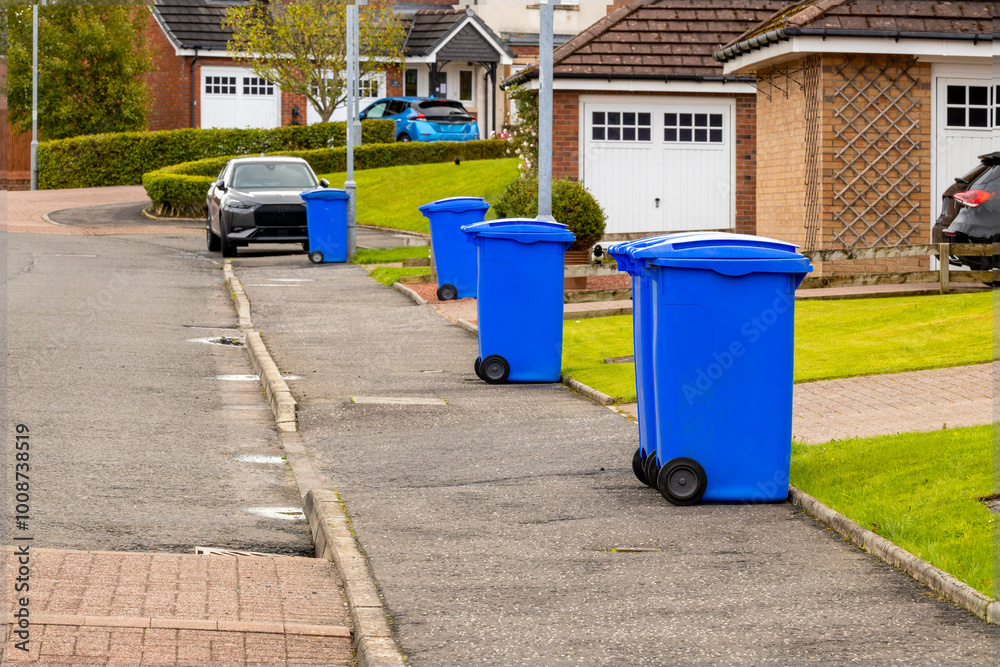 Blue waste recycling wheelie bins trash cans lined up in a row for collection on a residential street with parked cars Blue waste recycling wheelie bins trash cans lined up in a row for collection on a residential street with parked cars