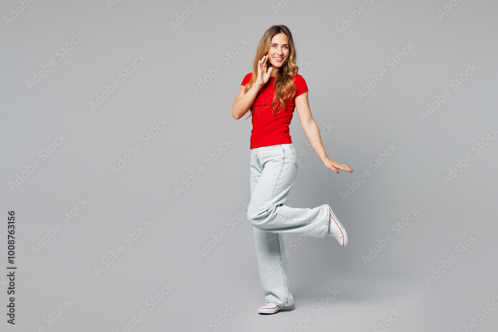 Full body side view young smiling happy cute woman she wear red t-shirt casual clothes put hand on face raise up leg look camera isolated on plain grey background studio portrait. Lifestyle concept. Full body side view young smiling happy cute woman she wear red t-shirt casual clothes put hand on face raise up leg look camera isolated on plain grey background studio portrait. Lifestyle concept.