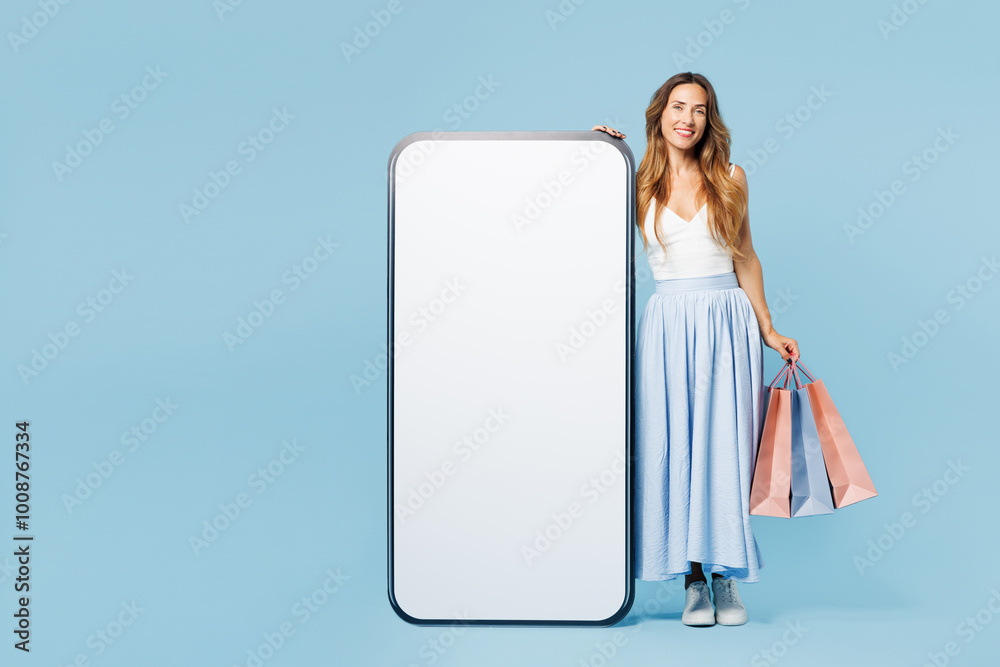 Full body young woman wear casual clothes hold shopping paper package bags stand near big huge blank screen area mobile cell phone isolated on plain blue background. Black Friday sale buy day concept. Full body young woman wear casual clothes hold shopping paper package bags stand near big huge blank screen area mobile cell phone isolated on plain blue background. Black Friday sale buy day concept.