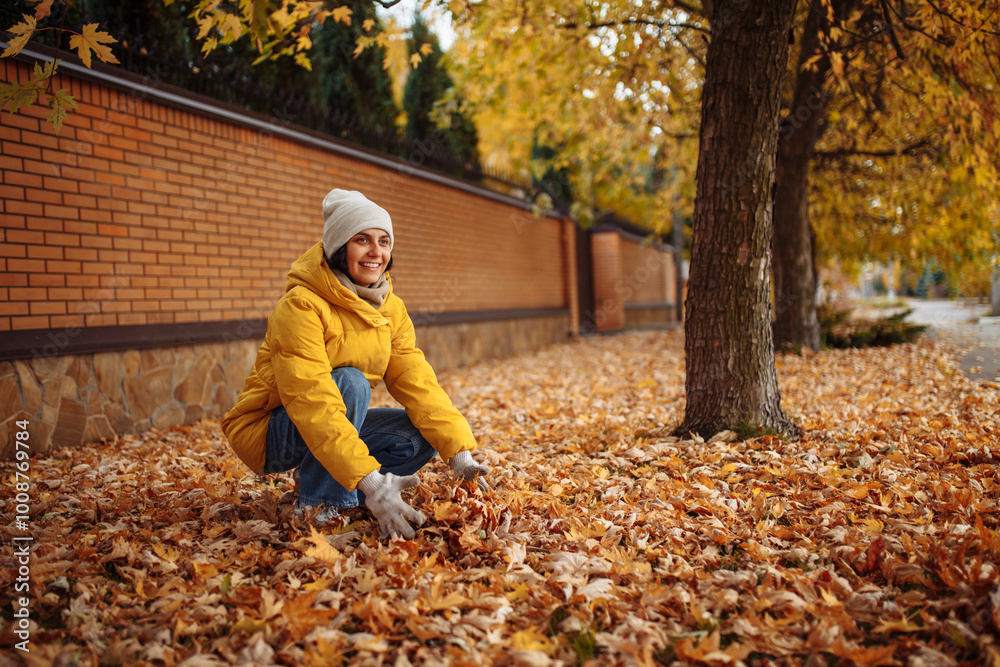 A woman in a yellow jacket and white gloves kneels, collecting vibrant autumn leaves. The ground is a colorful tapestry of orange, red, and yellow, embodying the beauty of fall. A woman in a yellow jacket and white gloves kneels, collecting vibrant autumn leaves. The ground is a colorful tapestry of orange, red, and yellow, embodying the beauty of fall.