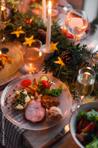 Christmas feast table with plates of food, glasses of wine and champagne, with festive decorations of pinecones, golden stars paper garland and long candles in evergreen foliage centerpieces