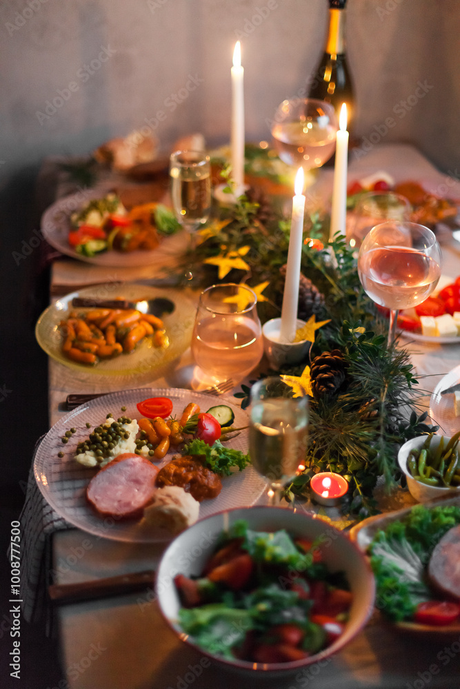 Fototapeta premium Christmas feast table with plates of food, glasses of wine and champagne, with festive decorations of pinecones, golden stars paper garland and long candles in evergreen foliage centerpieces