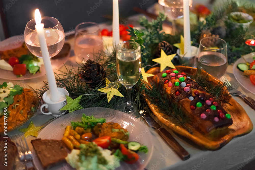 Fototapeta premium Christmas feast table with plates of food, glasses of wine and champagne, with festive decorations of pinecones, golden stars paper garland and long candles in evergreen foliage centerpieces