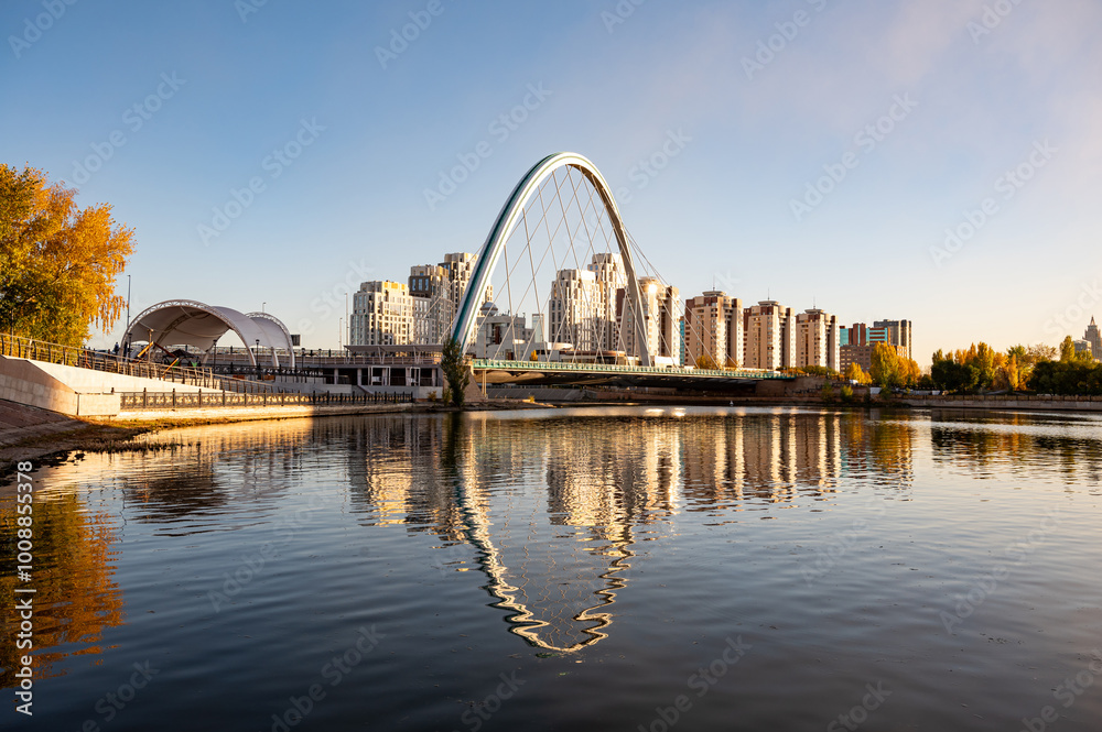 Fototapeta premium Karotkel Bridge over the Ishim River in Astana, Kazakhstan, with modern city buildings and autumn trees reflecting in the calm water under a clear sky