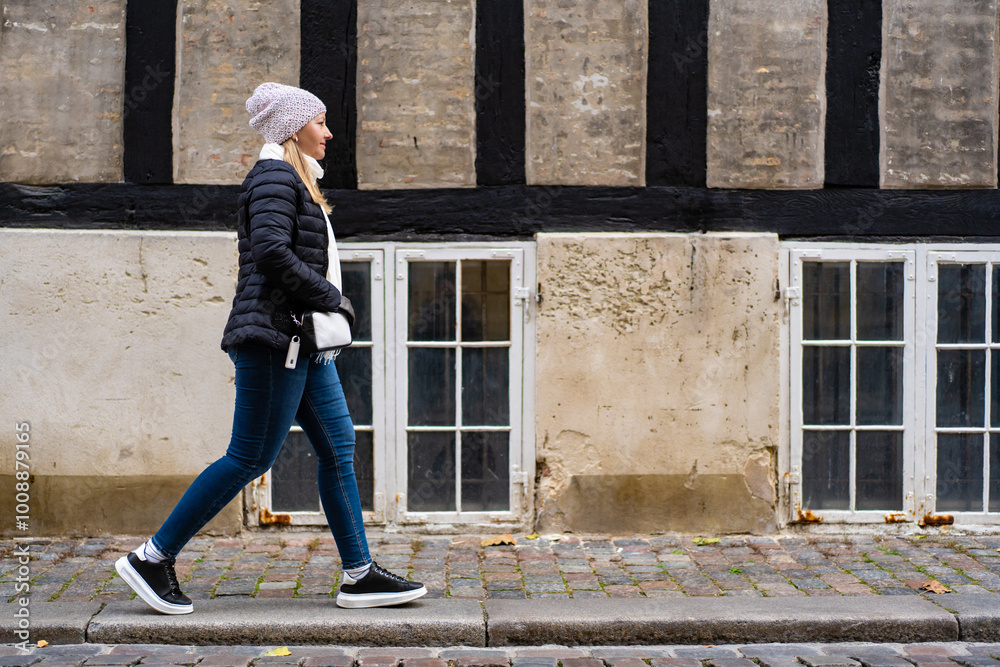 Visiting old city. Beautiful young woman in denim pants, black jacket and hat walking on sidewalk in old town against background of half-timbered wall. Side view. Timber framing architecture. Visiting old city. Beautiful young woman in denim pants, black jacket and hat walking on sidewalk in old town against background of half-timbered wall. Side view. Timber framing architecture.