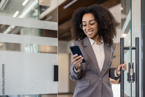 Smiling young African American business woman in a suit holds a phone in her hand and walks into the office opening the glass door