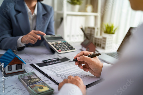 A man and a woman are sitting at a desk with a contract in front of them. The man is holding a pen and the woman is holding a calculator. Scene is serious and focused as they discuss the contract