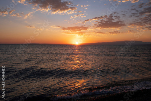 Dawn over the water of a lake or sea, the sun with its rays rises above the horizon and leaves a sunny path on the water and waves