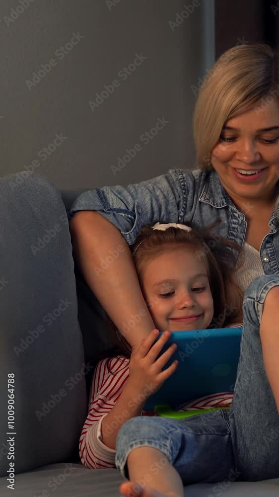 Mother and child watch tablet sitting on sofa at home in evening. Leisure. Happy family surfing internet. Social media, computer games. Vertical shot Mother and child watch tablet sitting on sofa at home in evening. Leisure. Happy family surfing internet. Social media, computer games. Vertical shot
