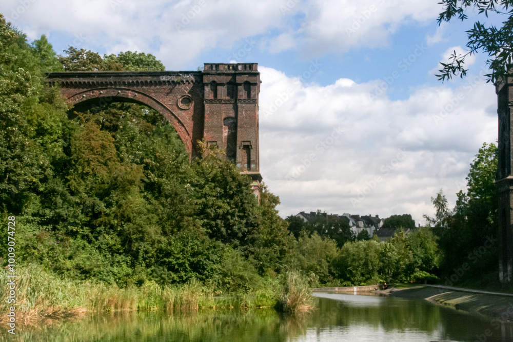 Fototapeta premium Old viaduct bridge covered with green trees and bushes next to a pond.