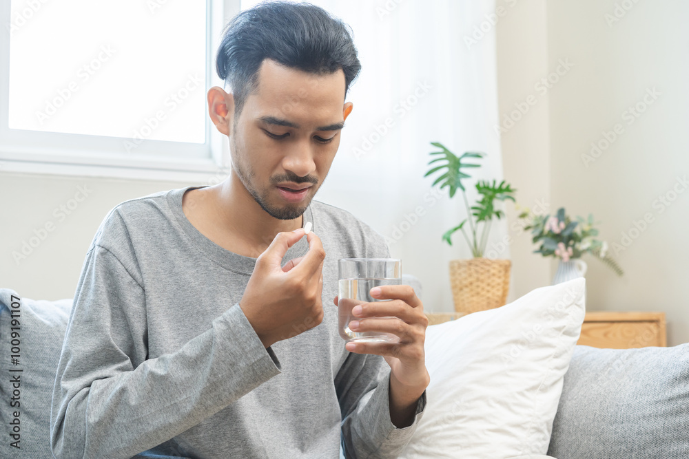 Sick asian young man, male holding tablet pill on hand, painkiller medicine from fever, stomach pain, headache for treatment, take drug or vitamin with glass of water on at home. Health care concept. Sick asian young man, male holding tablet pill on hand, painkiller medicine from fever, stomach pain, headache for treatment, take drug or vitamin with glass of water on at home. Health care concept.