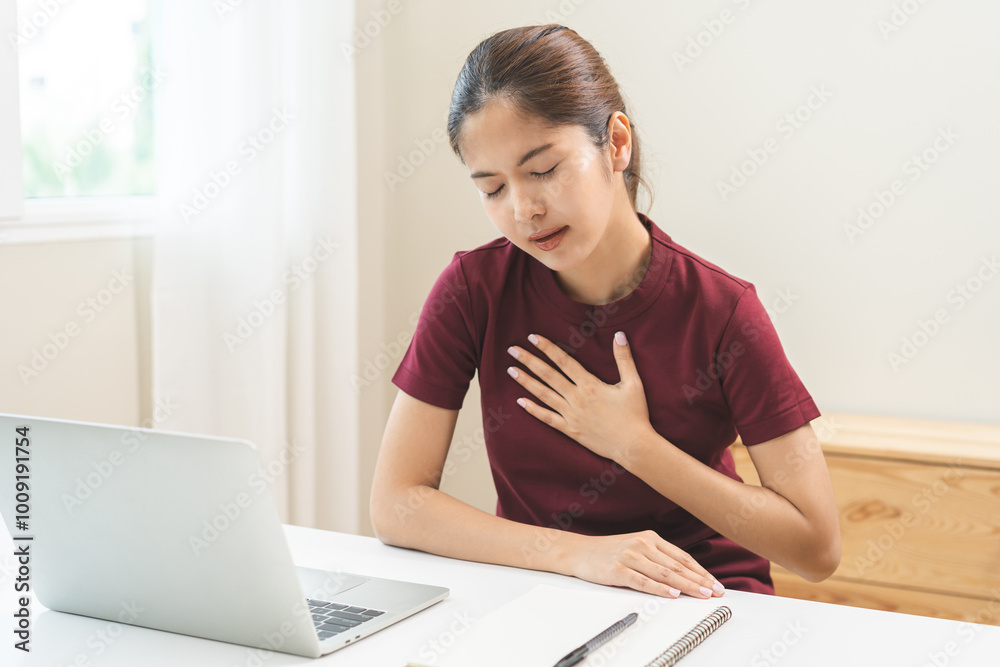 Heart attack, health care concept, suffering acute cardiac asian young woman hand touching on her chest pain, breathing problem, asthma attack, pressure while sit work with computer, laptop at home. Heart attack, health care concept, suffering acute cardiac asian young woman hand touching on her chest pain, breathing problem, asthma attack, pressure while sit work with computer, laptop at home.