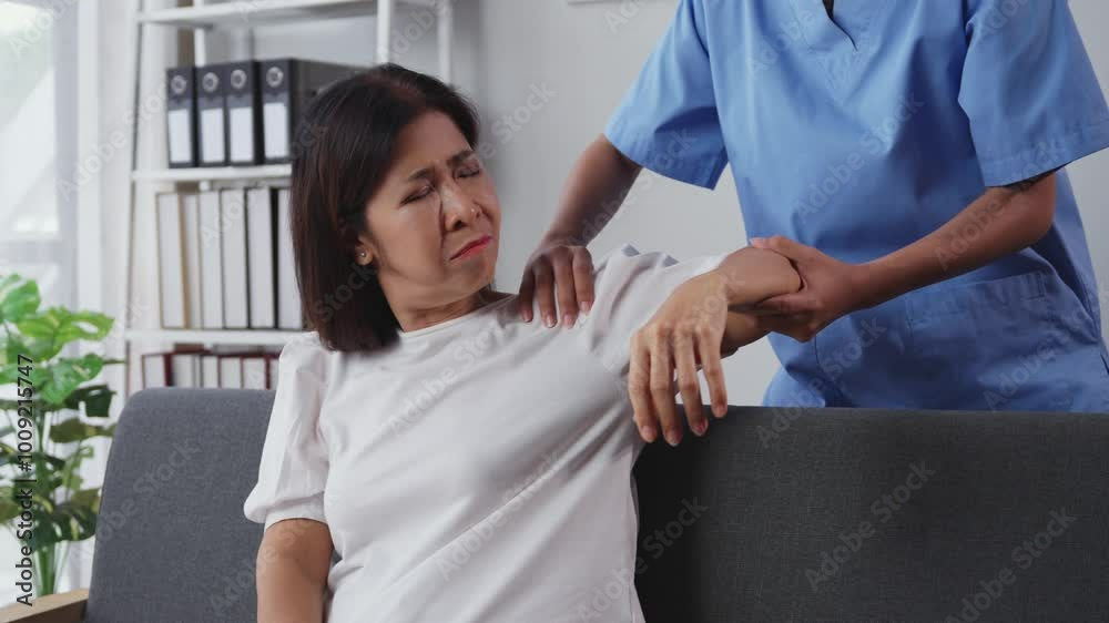 Nurse wearing blue scrubs gently massages the arm of an elderly woman sitting on a sofa who is wincing in pain Nurse wearing blue scrubs gently massages the arm of an elderly woman sitting on a sofa who is wincing in pain