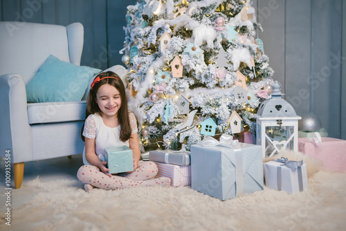 Laughing small girl sitting near white Christmas tree with present boxes. Grey and blue colors modern interior. Holding gift waiting to open it. New year eve family winter holidays mood