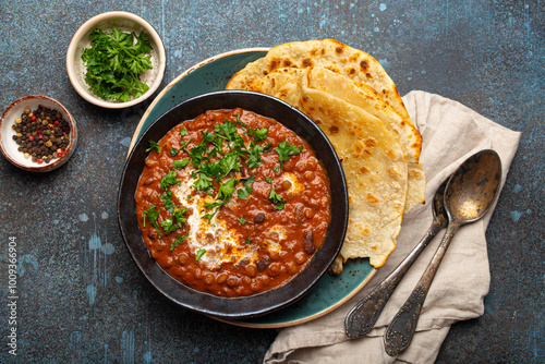 Traditional Indian Punjabi dish Dal makhani with lentils and beans in black bowl served with naan flat bread, fresh cilantro and two spoons on blue concrete rustic table top view.