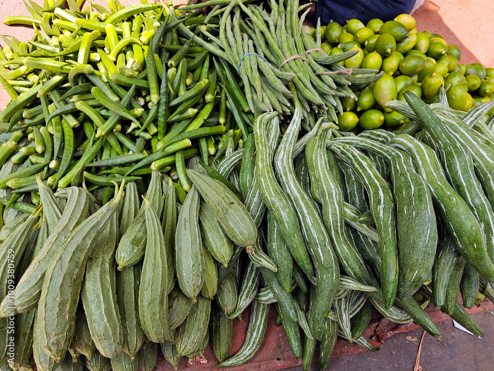 variety of fresh green vegetables displayed at a market. okra, trichosanthes, abelmoschus esculentus, long beans, ridge gourd, and limes, all arranged neatly.