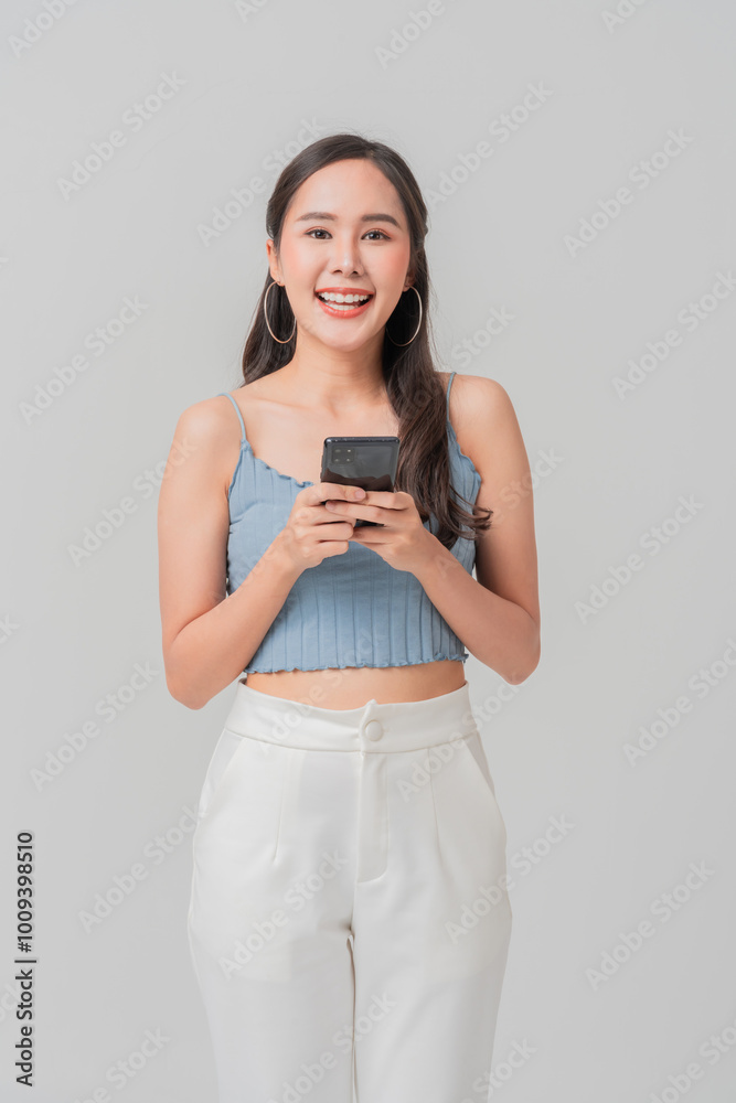 Happy young woman holding smartphone, wearing light blue crop top and white pants, exuding confidence and joy. She stands against neutral background, ready to connect