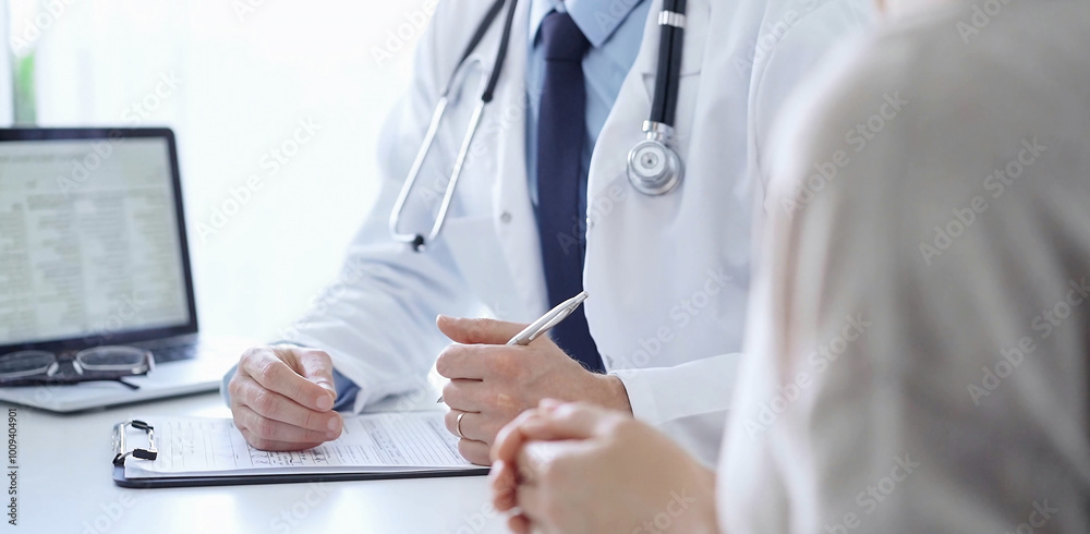 A doctor and a patient. The physician, wearing a white medical coat over a blue shirt and tie, is filling out a medical record form during a consultation in the clinic. Medical service A doctor and a patient. The physician, wearing a white medical coat over a blue shirt and tie, is filling out a medical record form during a consultation in the clinic. Medical service