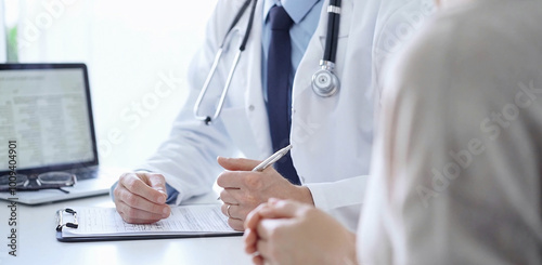 A doctor and a patient. The physician, wearing a white medical coat over a blue shirt and tie, is filling out a medical record form during a consultation in the clinic. Medical service