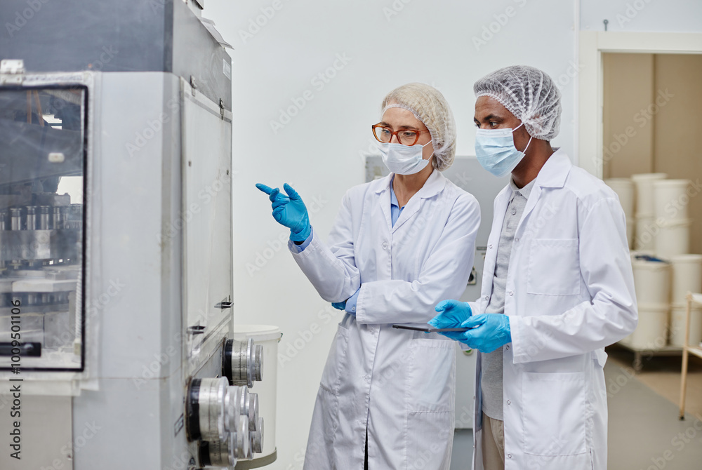 Fototapeta premium Medium shot of middle aged female process technician in lab coat pointing at machine, while inspecting drug production with African American male colleague at pharmaceutical manufacturing plant