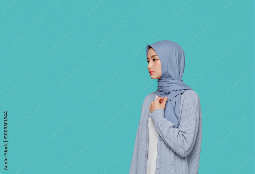 Fototapeta premium A woman in a soft blue hijab and matching cardigan gazes thoughtfully to the side, standing against a turquoise backdrop. Her calm demeanor reflects simplicity and elegance. Generative AI