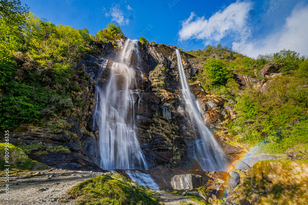 Fototapeta premium A majestic double waterfall plunges down rugged cliffs, with a rainbow shimmering at the bottom. Bright foliage and clear skies enhance the scenic beauty of this natural wonder