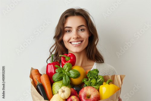 A smiling woman holding an open brown paper bag full of colorful vegetables and fruits.