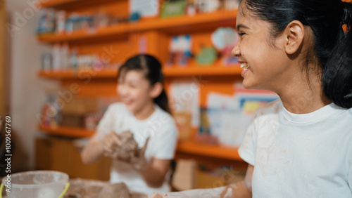 Young happy caucasian girl modeling cup with friend at pottery workshop. Smart asian girl smash dough while attend in art lesson when friend doing creative activity. Blurred background. Edification.