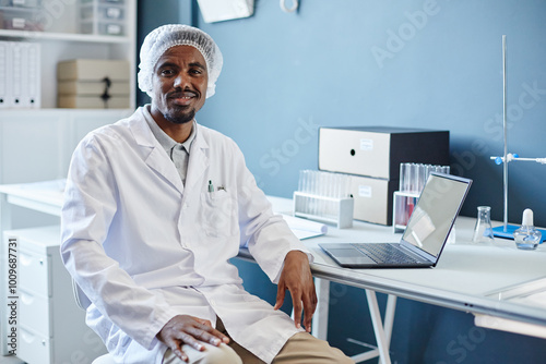 Medium portrait shot of African American male lab chemist in medical hairnet smiling at camera while sitting at working table in laboratory of scientific research service, copy space