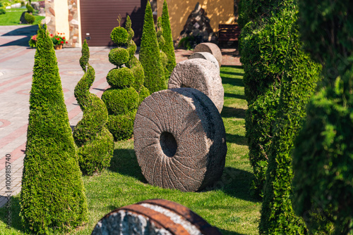 Well-kept garden with a variety of topiary shrubs and a decorative stone mill wheel. With a brick pathway leading into the center of the garden. The trees and shrubs have been carefully shaped. photo