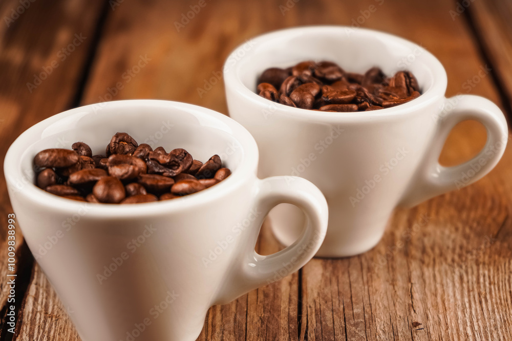 Two white ceramic cups with roasted coffee beans inside on brown wooden table. Coffee shop, caffeine, roast concept. Roasted beans for mass market sale, horizontal photo. Two white ceramic cups with roasted coffee beans inside on brown wooden table. Coffee shop, caffeine, roast concept. Roasted beans for mass market sale, horizontal photo.