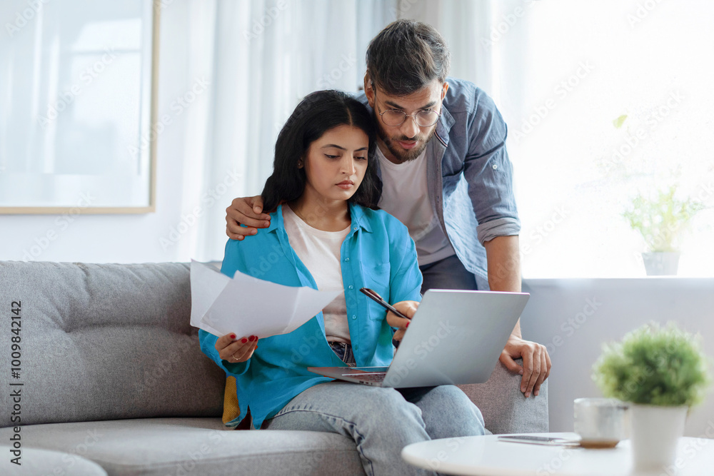 Household papers problem. Indian couple looking at laptop and holding documents, reading bills and correspondence, home interior Household papers problem. Indian couple looking at laptop and holding documents, reading bills and correspondence, home interior