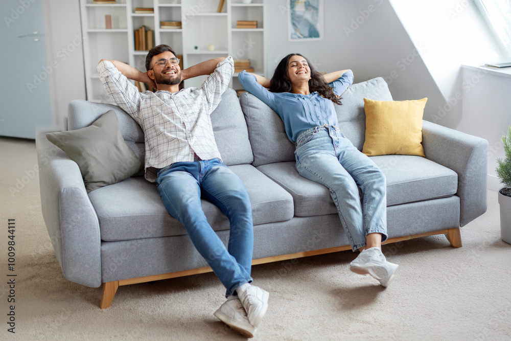 Joyful hindu couple lounges on sofa, hands behind their heads, enjoying leisurely moment in living room interior at home setting Joyful hindu couple lounges on sofa, hands behind their heads, enjoying leisurely moment in living room interior at home setting