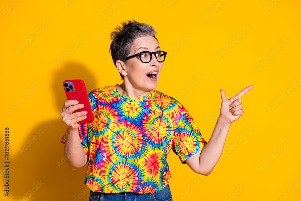 Photo of nice aged lady hold phone point finger empty space wear t-shirt isolated on yellow color background Photo of nice aged lady hold phone point finger empty space wear t-shirt isolated on yellow color background