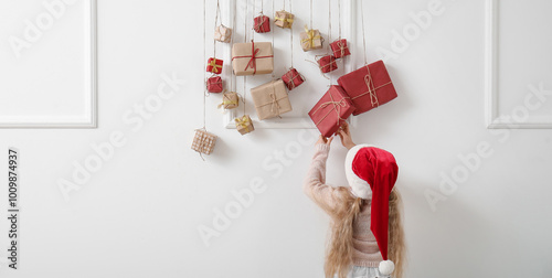 Cute little girl in Santa hat and Christmas advent calendar hanging on light wall