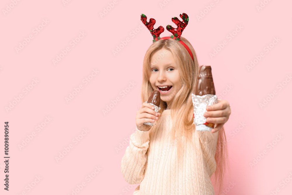 Φωτογραφία Cute little girl in reindeer horns eating chocolate Santa Claus on pink backgrou