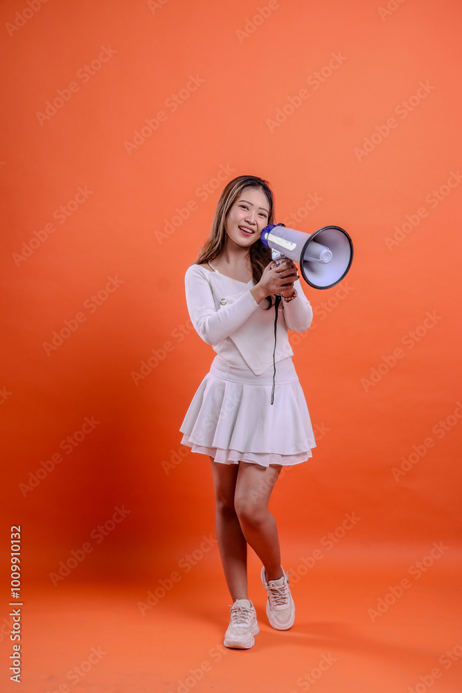 Fototapeta premium full length beautiful asian girl white top standing to camera screaming cheerful both hands holding megaphone loudspeaker isolated orange background. for technology, advertising, broadcast, promotion
