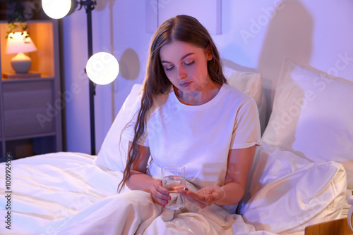 Young woman with glass of water taking sleeping pills in bedroom at night photo