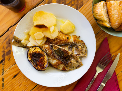 There is plate with lunch on simple wooden table. Dish is made from fried fish carcass of lean dorado, baked potato vegetables and several round pieces of eggplant photo