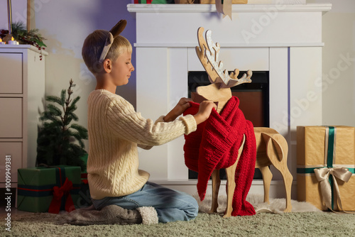Little boy wrapping wooden reindeer with scarf at home on Christmas eve
