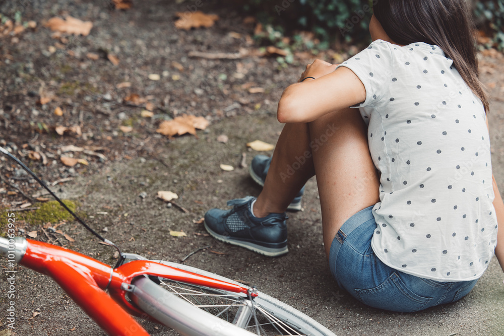 Obraz premium Young caucasian woman riding a red bicycle, captured moments before and after an accident outdoors. Accident with a bike, hurt her knee after she fell on the road on the way home.