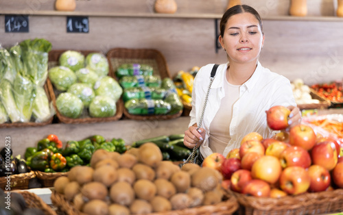 Portrait of positive interested young woman buying fruits in greengrocery store, choosing ripe apples arranged in woven box on produce display.. photo