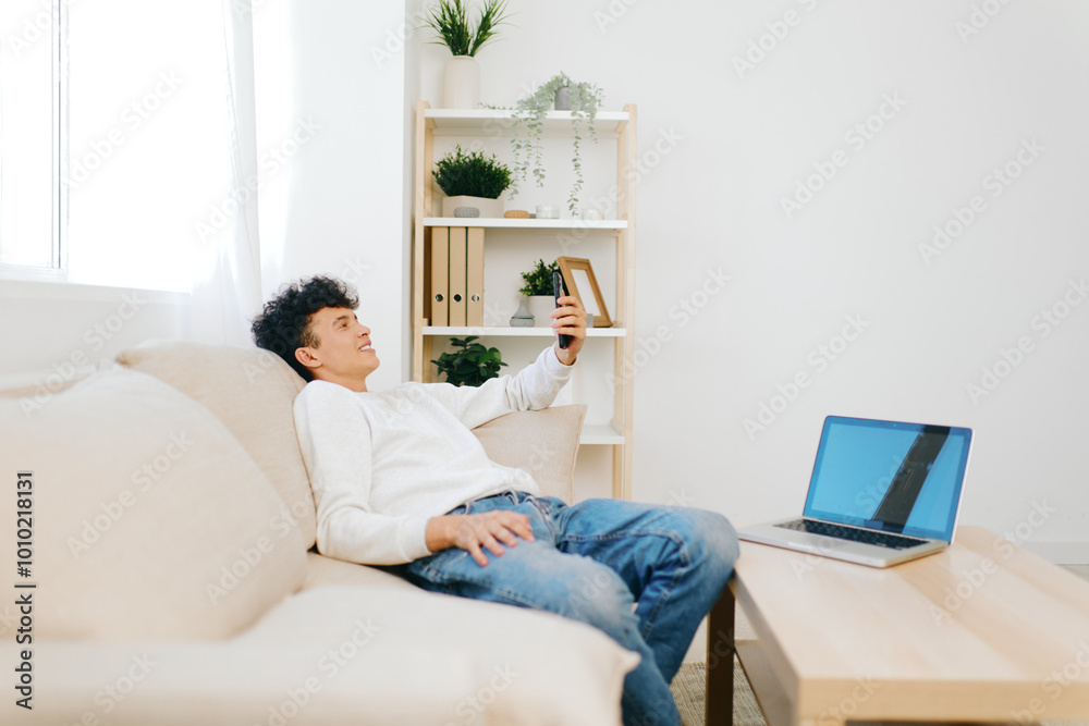 © SHOTPRIME STUDIO - Young man using smartphone while relaxing on sofa in minimalistic home interior with laptop on table, bright and airy space designed for comfort and productivity © SHOTPRIME STUDIO - Young man using smartphone while relaxing on sofa in minimalistic home interior with laptop on table, bright and airy space designed for comfort and productivity